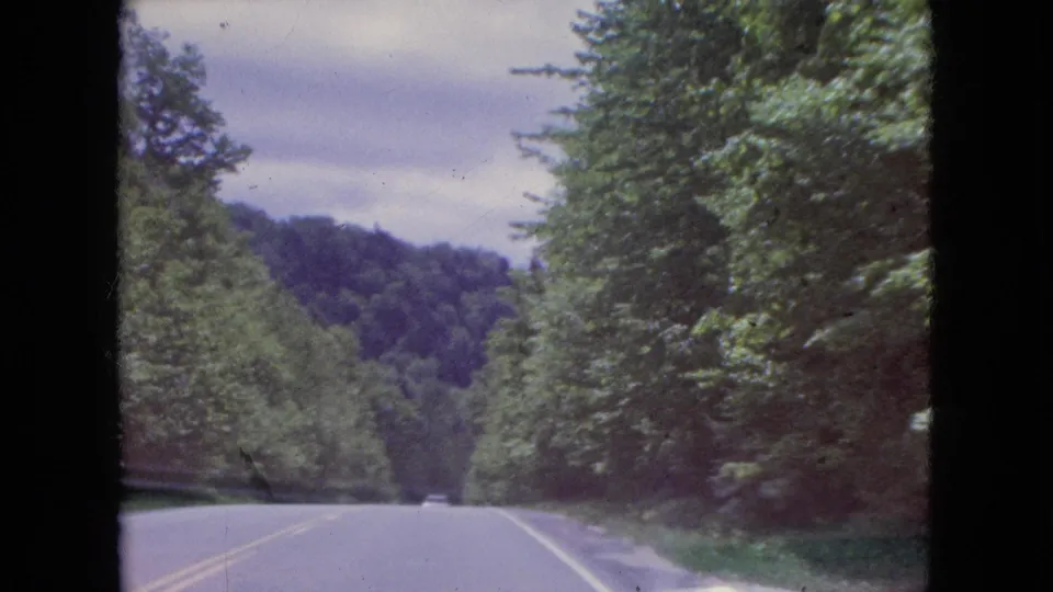 Passenger-view 1964 8mm frame looking down a tree-lined two-lane highway toward a wooded hillside in the Niagara Falls, New York area.