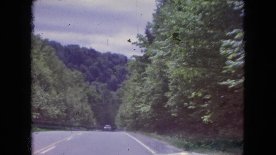 1964 8mm home-movie frame showing a two-lane road through dense green trees and forested hills near Niagara Falls, New York, with a distant car ahead.