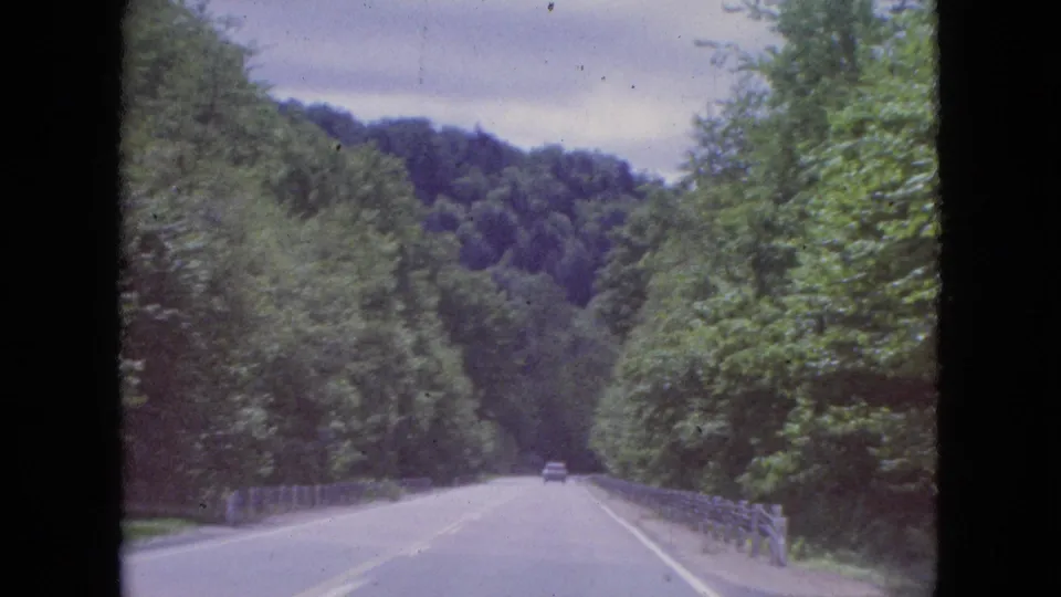 A moving-vehicle view of a two-lane road with roadside guardrails, dense trees, and forested hills in 1964 home-movie footage from the Niagara Falls, New York area.