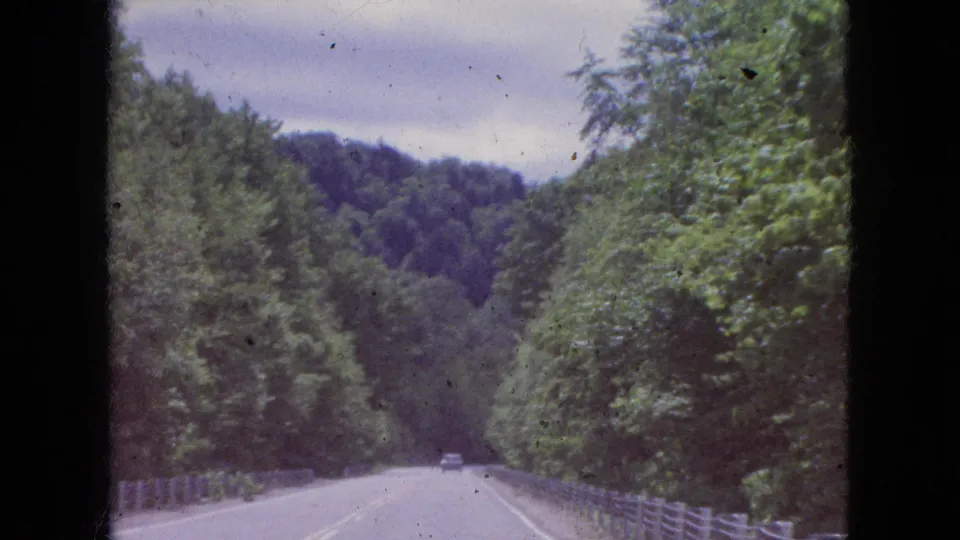 1964 8mm travel frame looking down a tree-lined two-lane road with wooden guardrails, a distant vehicle, and wooded hills in the background.