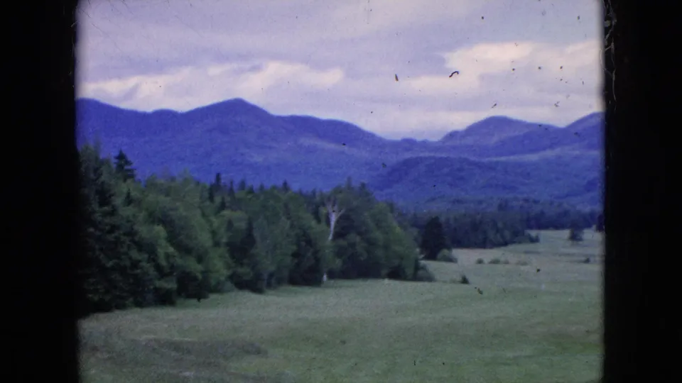 1964 8mm home-movie frame of an open green golf-course landscape in the United States, with a tree line, distant mountains, cloudy sky, and dark film edges.