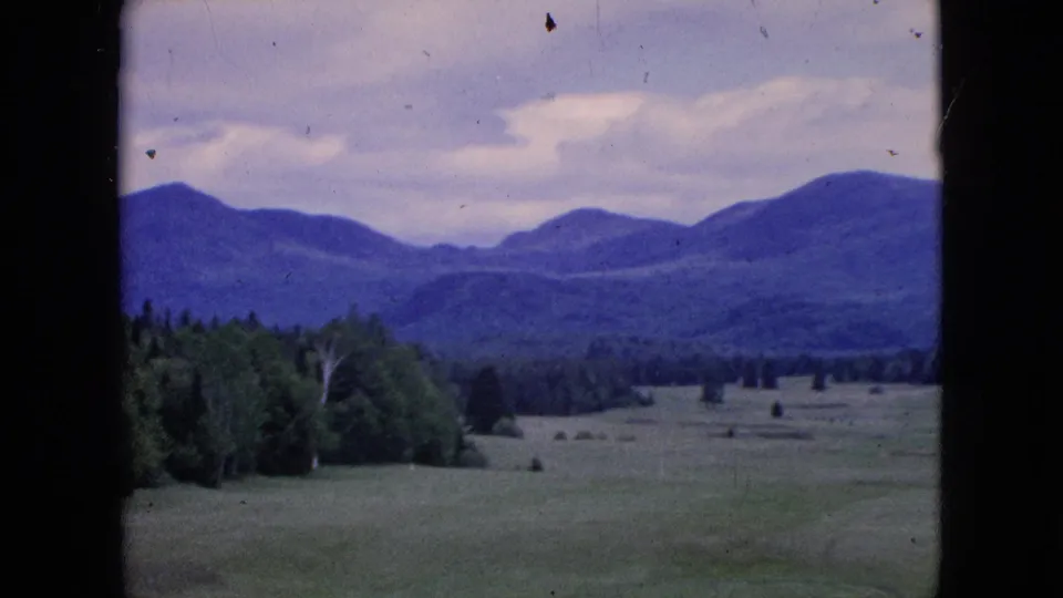 Panned 1964 home-movie view across a green recreational landscape, showing trees, distant blue mountains, overcast sky, and visible 8mm frame edges.