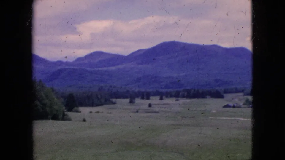 1964 8mm frame of a broad green course or fairway with mountains beyond, a wooded edge, cloudy sky, and small distant structures visible near the horizon.