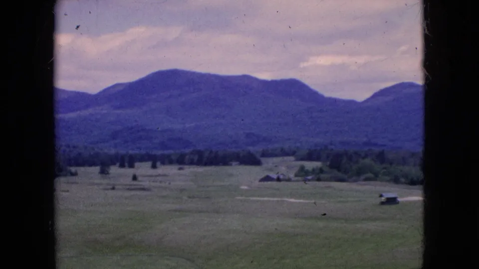 Final panned 1964 home-movie frame showing a green recreational landscape, distant mountains, a tree line, small buildings, and a pale path or road under cloud cover.