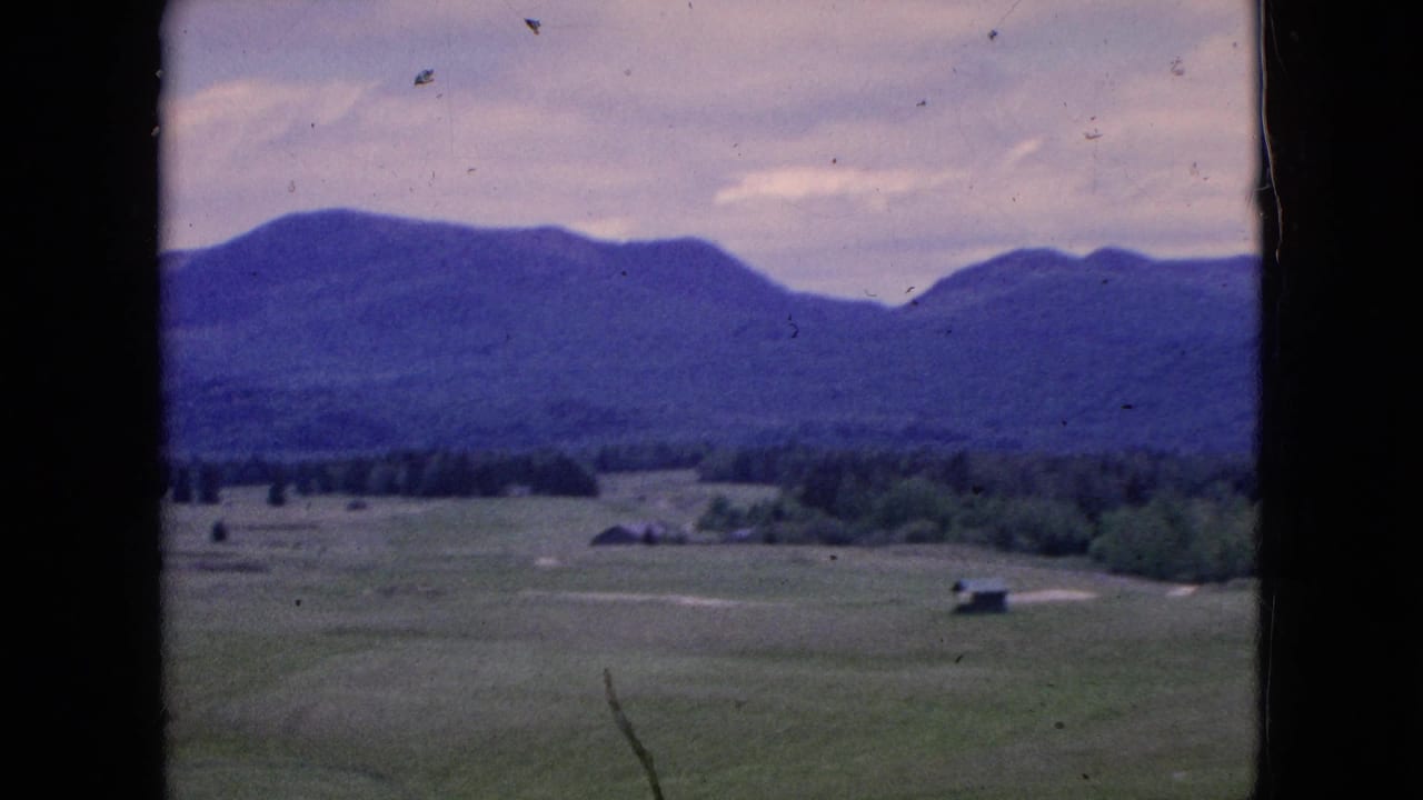 1964 8mm home-movie view of green fields, small distant buildings, treeline, and blue-toned hills framed by the dark edge of a moving car window.
