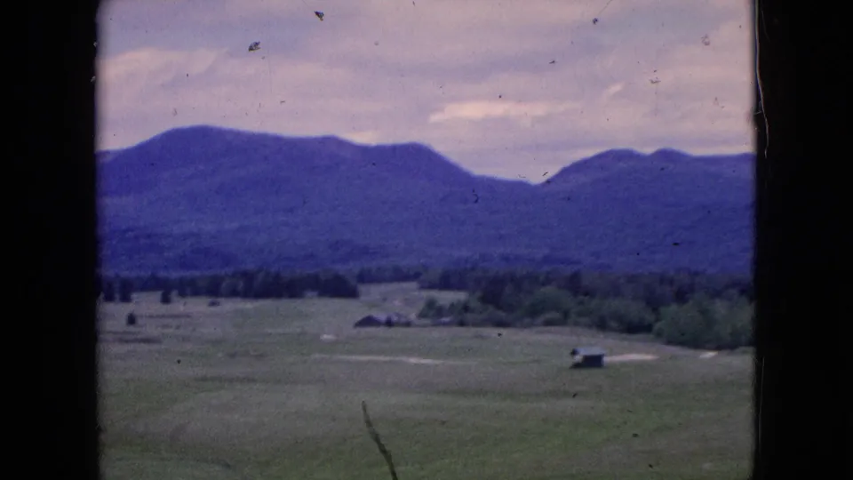1964 8mm home-movie view of green fields, small distant buildings, treeline, and blue-toned hills framed by the dark edge of a moving car window.