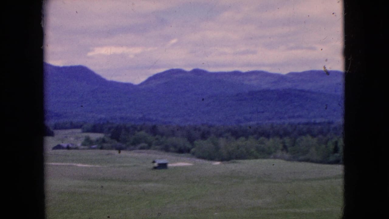 1964 8mm travel footage showing open green fields, a small distant structure, dark treeline, and hazy blue hills beneath a soft overcast sky.