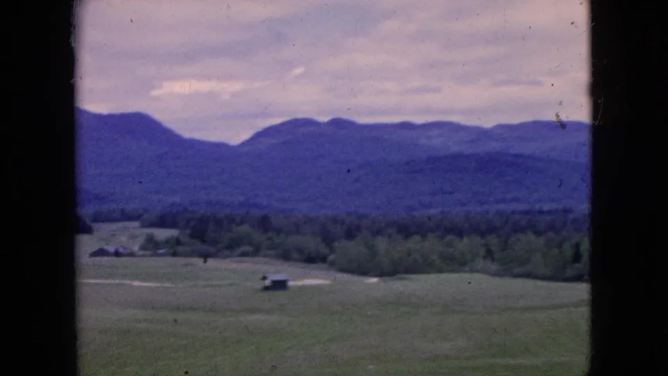 1964 8mm travel footage showing open green fields, a small distant structure, dark treeline, and hazy blue hills beneath a soft overcast sky.