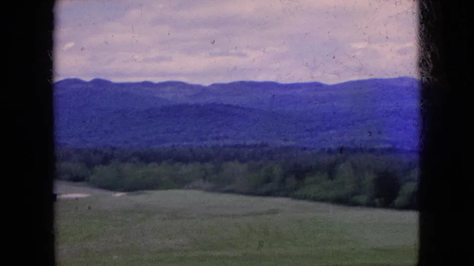 Soft 8mm frame from a moving vehicle, with foreground fields, a dark treeline, and distant blue hills partially blurred by motion and film texture.