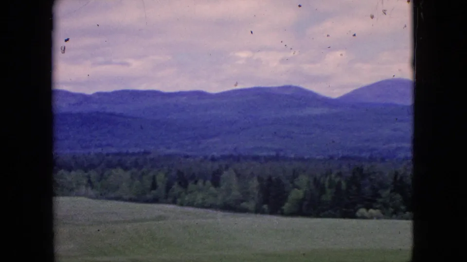 1964 8mm landscape frame with a green field in front, dense trees across the middle distance, and blue-toned hills under a cloudy sky.