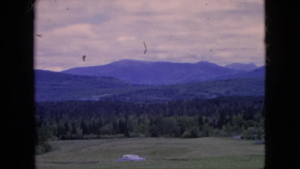 1964 8mm home-movie frame showing a pale classic car in a field, with dark window edges, tree-covered hills, and distant mountains under a cloudy sky.