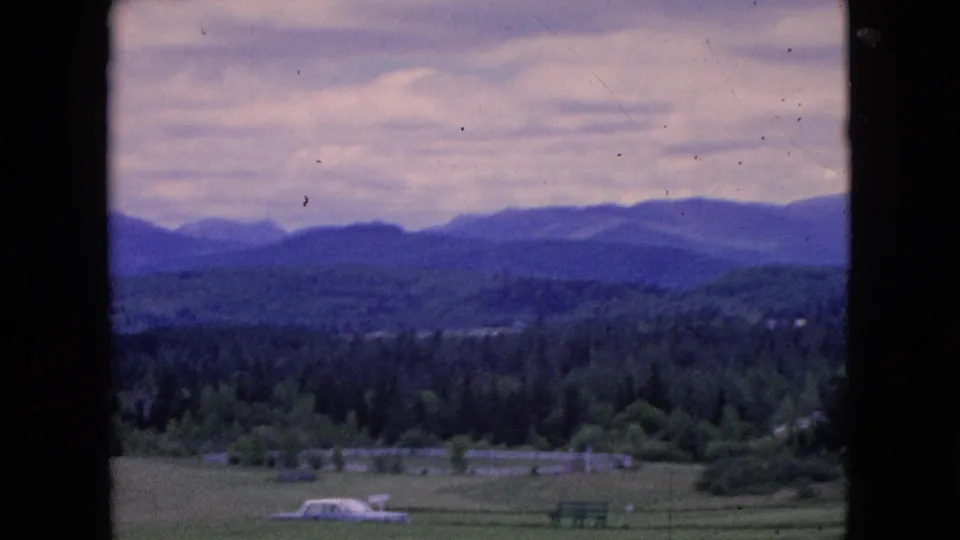 1964 8mm frame showing a pale car near the bottom edge of a rural field, with scattered structures, forest, hills, and distant mountain ridges beneath a cloudy sky.