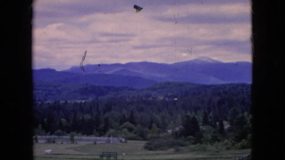 1964 home-movie frame looking across a rural field and tree line toward blue mountain ridges, with small structures and visible film dust in the sky.