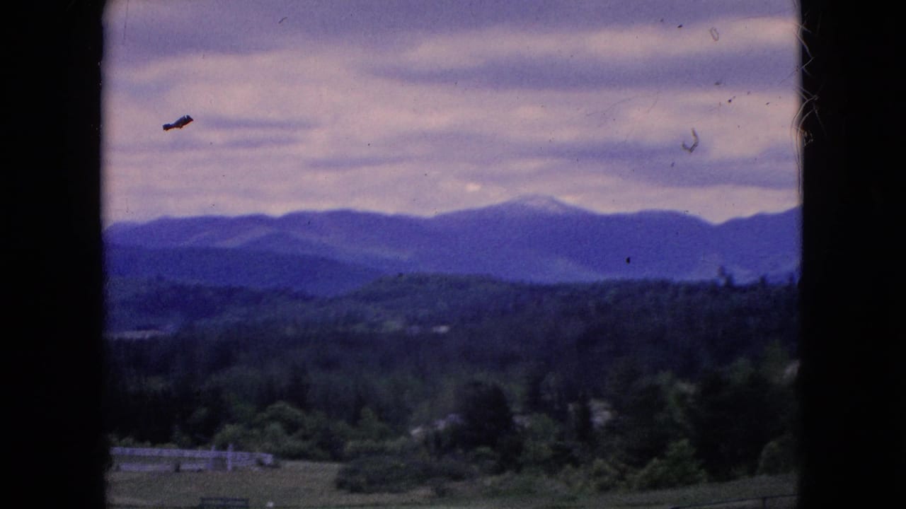 1964 home-movie frame from the Niagara Falls, New York metadata showing wooded countryside, distant blue-purple mountains, fields, fencing, and an overcast sky seen through a dark window frame.