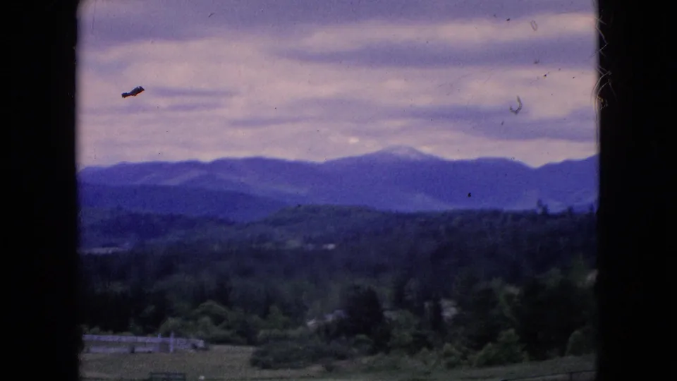 1964 home-movie frame from the Niagara Falls, New York metadata showing wooded countryside, distant blue-purple mountains, fields, fencing, and an overcast sky seen through a dark window frame.