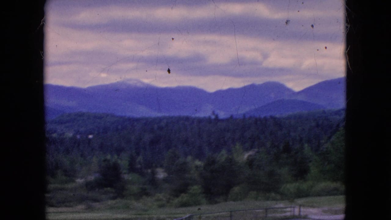 Window-framed 8mm view of layered mountains, dense tree cover, low fields, and a gray-purple overcast sky in footage identified as 1964 Niagara Falls, New York.