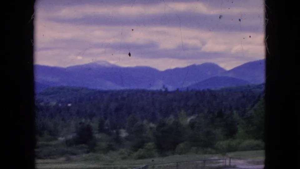 Window-framed 8mm view of layered mountains, dense tree cover, low fields, and a gray-purple overcast sky in footage identified as 1964 Niagara Falls, New York.