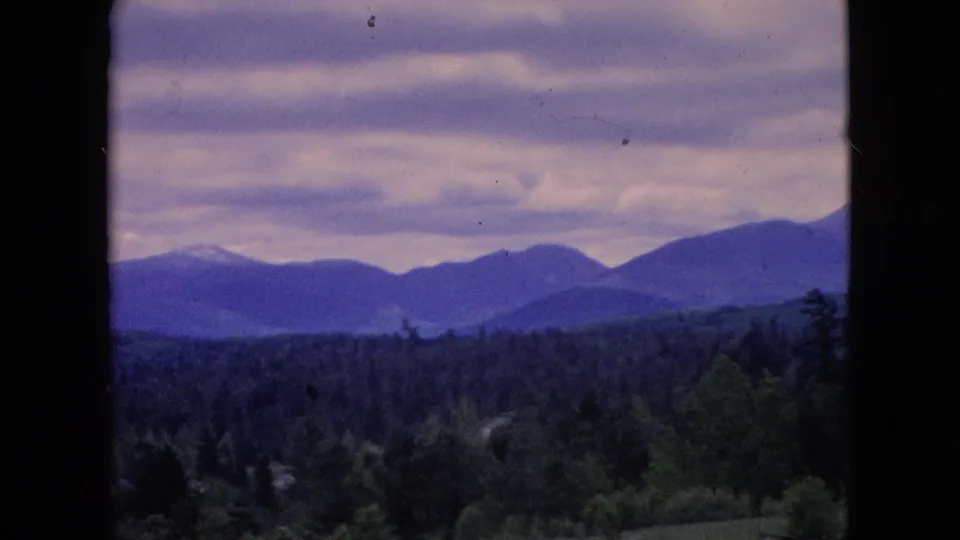 Archival 8mm frame showing a soft, purple-toned countryside view with distant mountains, dark tree line, green fields, and overcast clouds, consistent with the 1964 travel metadata.