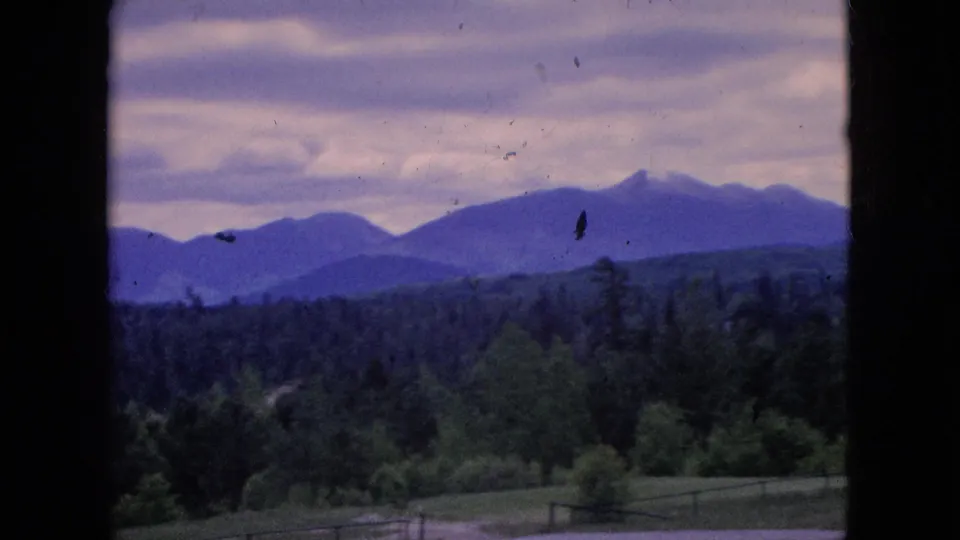 Final frame of the short 8mm landscape view, with purple-blue mountains, wooded hills, open foreground fields, a fence line, cloudy sky, and visible archival dust and scratches.