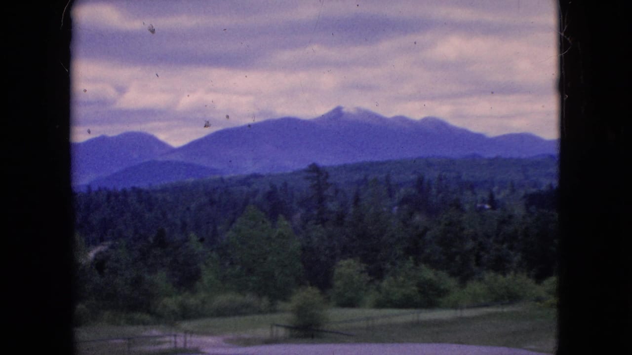 1964 8mm home-movie frame of a green mountain valley viewed through dark vertical window or doorway framing, with distant blue-purple ridges under a cloudy sky.
