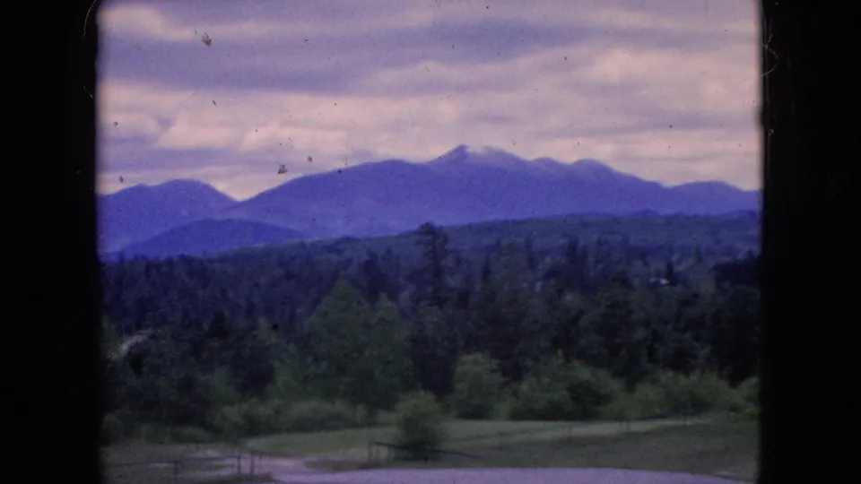 1964 8mm home-movie frame of a green mountain valley viewed through dark vertical window or doorway framing, with distant blue-purple ridges under a cloudy sky.