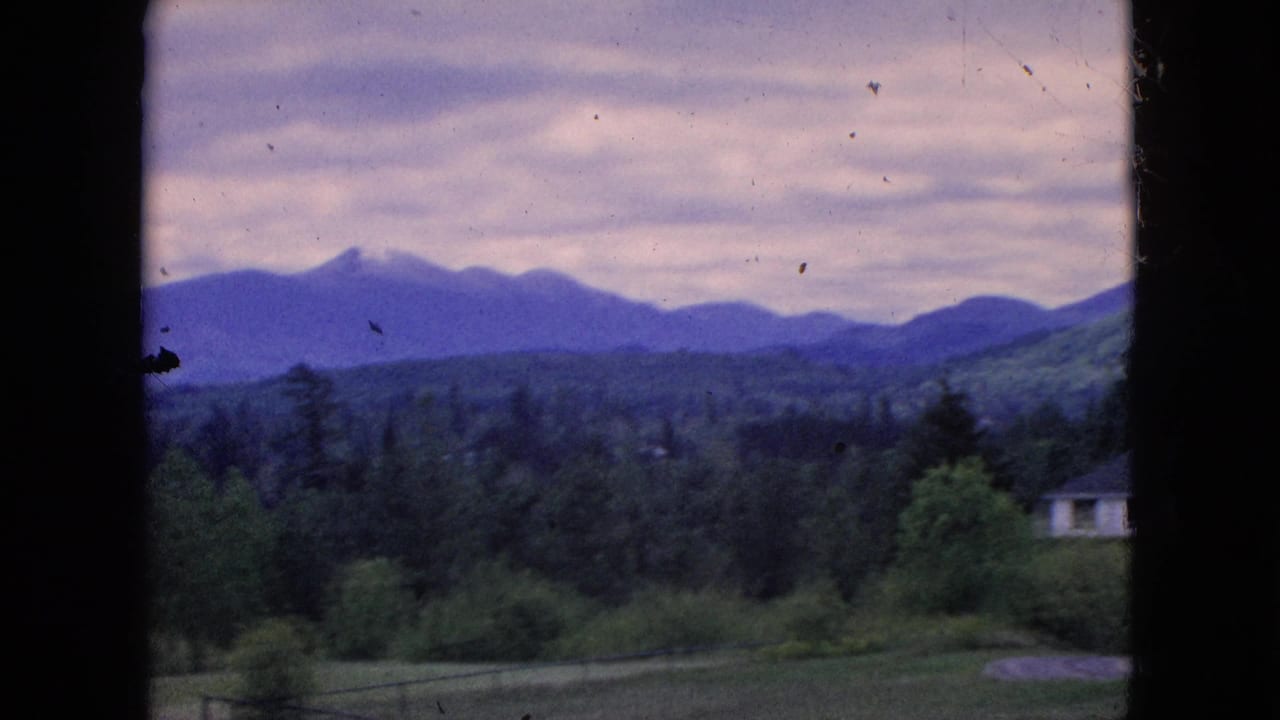1964 8mm frame showing layered mountains and a forested valley through dark foreground framing, with a small structure barely visible at the far right edge.