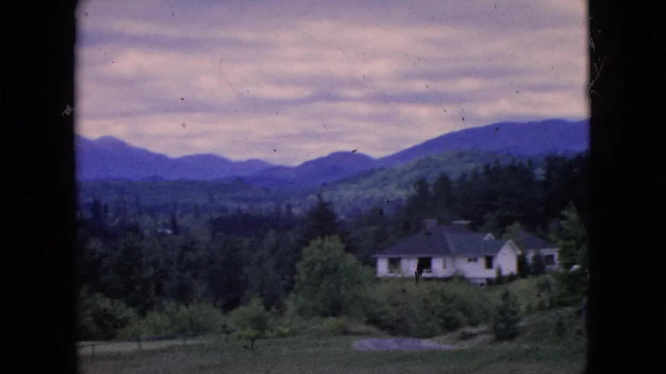 1964 8mm home-movie frame with a light-colored house and dark roof set among trees below distant mountain ridges in a green valley.