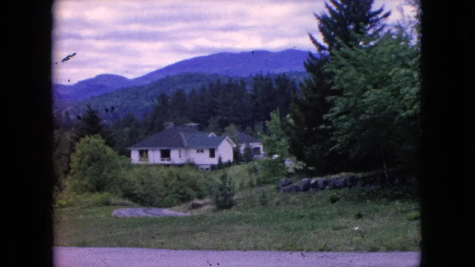 1964 8mm frame showing a light-colored house near a tall evergreen, shrubs, rocks, and a blue-purple mountain slope in the background.