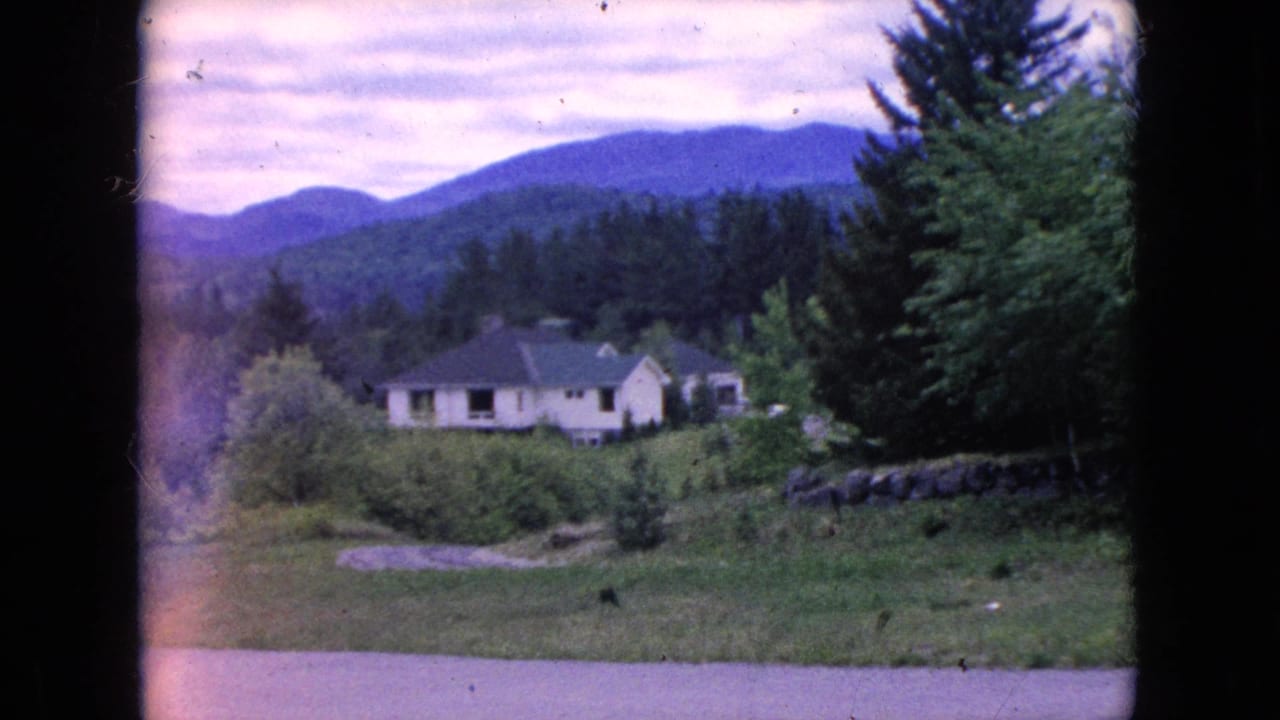 1964 home-movie frame of a small light-colored rural house in the Niagara Falls, New York area, seen across grass with evergreen trees and low mountains behind it.