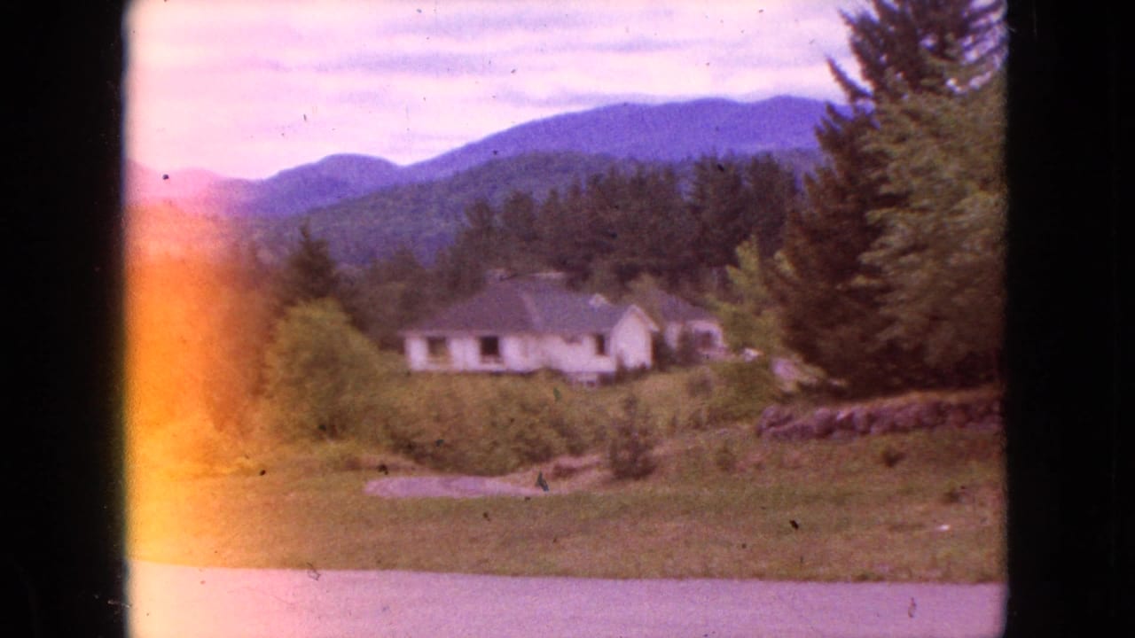 1964 8mm frame showing a distant rural house near Niagara Falls, New York, with evergreens, low mountains, open grass, and a warm exposure flare along the frame edge.