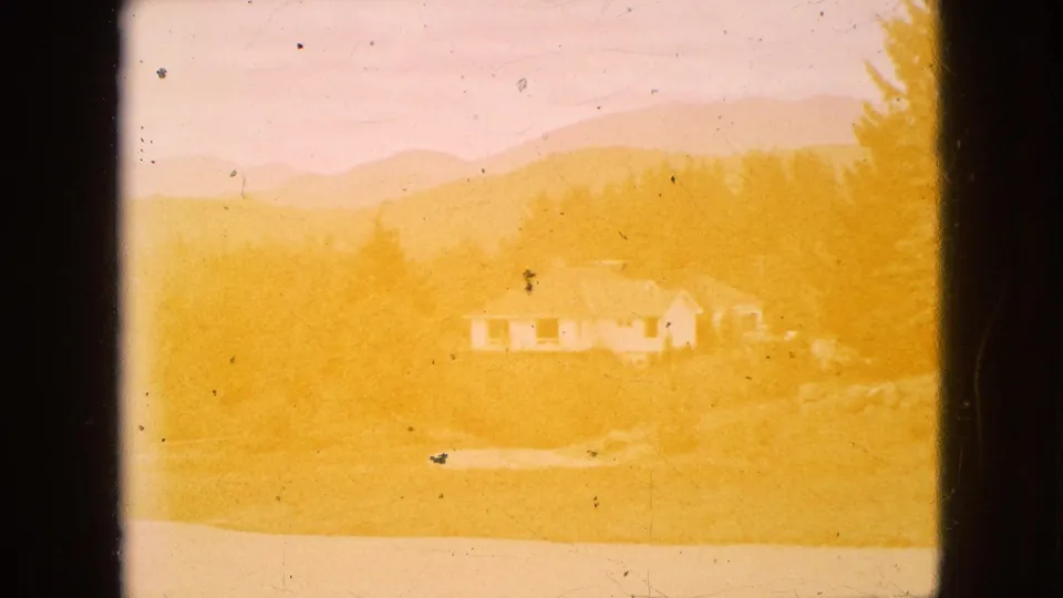 Overexposed 1964 home-movie frame of the rural house and hillside, with the scene partly washed by yellow light while trees and mountains remain faintly visible.