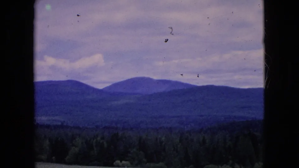 1964 8mm home-movie frame with layered blue-toned ridges, a dark tree line, open countryside, and a pale sky with light clouds.