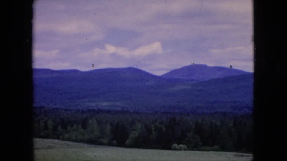 1964 8mm home-movie frame showing a rolling green field in the foreground, dense trees, distant rounded hills, and a muted purple-blue sky.