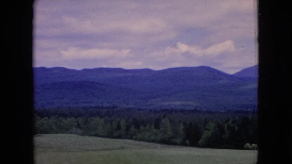 1964 8mm home-movie frame of an open rural field, dark conifer forest, and a long low hill range beneath a pale clouded sky.