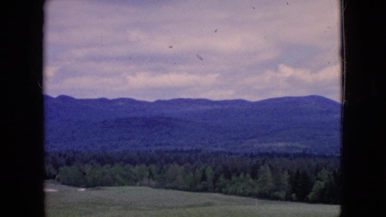 1964 8mm home-movie frame of a rural landscape in the United States, with green fields, a tree line, distant hills, and cloudy sky seen through dark frame edges.