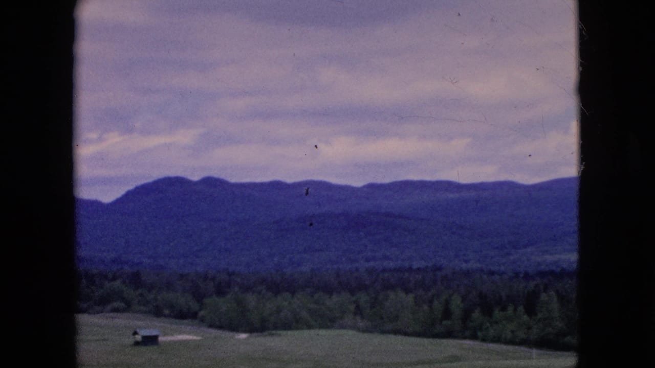 1964 8mm home-movie frame showing an open rural field with a small shed, dense trees, and distant blue hills under a soft cloudy sky.