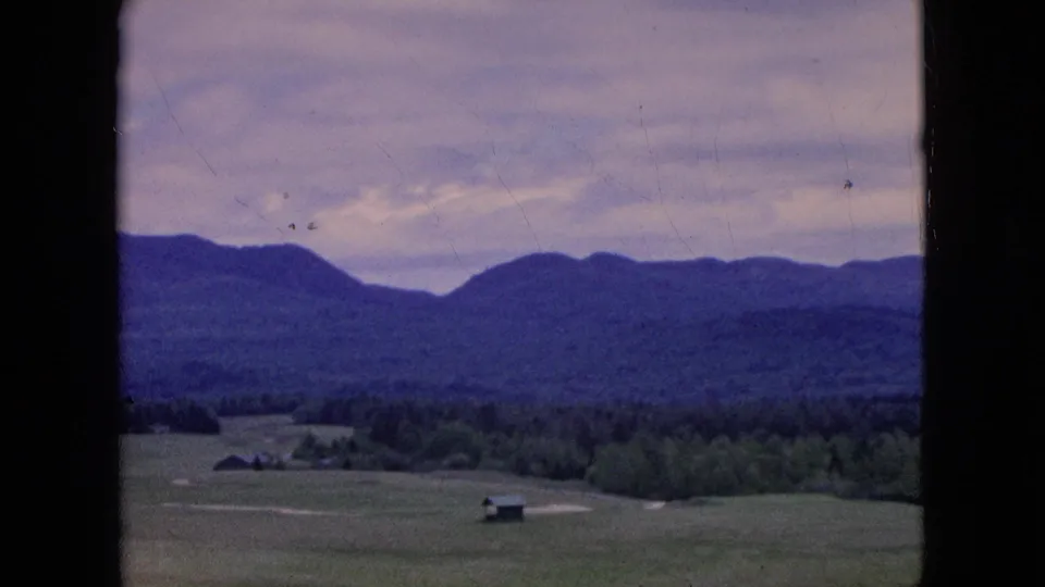 1964 8mm home-movie frame of a broad rural valley, with open fields, scattered low buildings, dense trees, and distant hills under a cloudy sky.