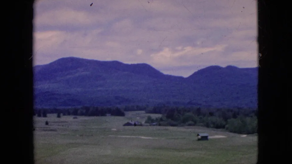 1964 8mm home-movie frame showing fields and small rural buildings in front of a wooded tree line and blue hills beneath a cloudy sky.