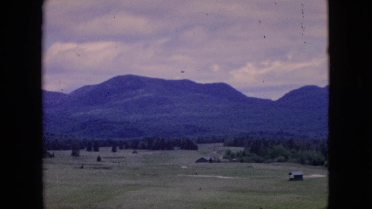 1964 8mm home-movie frame showing open rural fields, small distant structures, a dark tree line, and a broad mountain ridge beneath a muted sky.