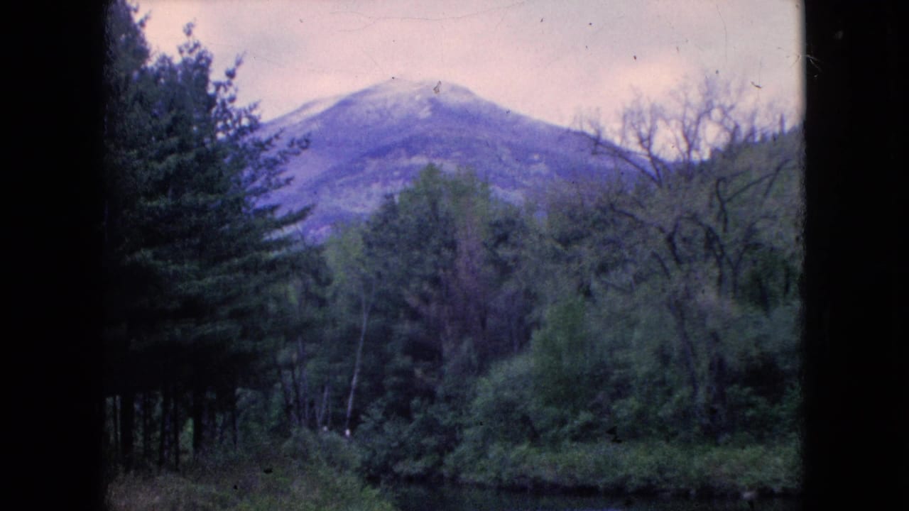 1960s 8mm home-movie frame showing a wooded river valley in the United States with a distant rounded mountain, pale sky, and dark film borders.