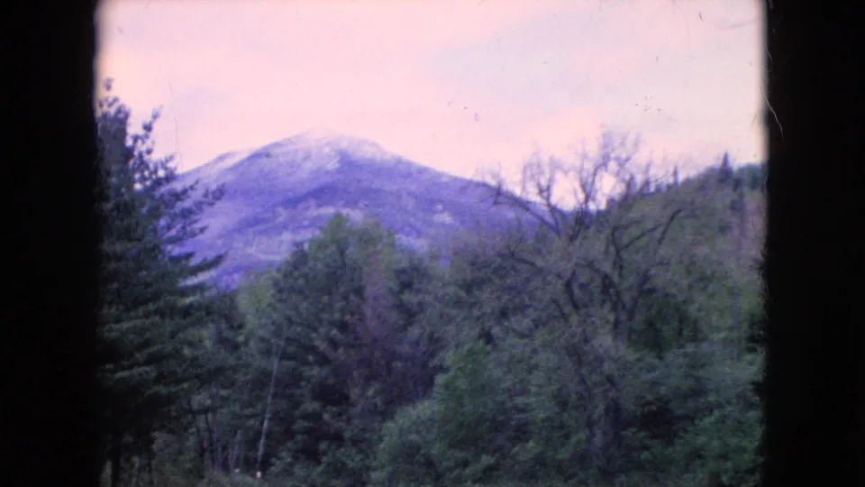 1960s 8mm home-movie frame with a rounded mountain in the distance behind dense forest and a partially visible river valley in the United States.