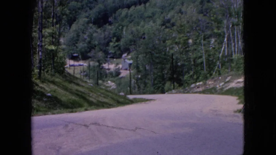 1964 8mm travel frame showing a curving light-colored road through a wooded mountain area, with dense trees, roadside slope, and small distant structures or objects that are not clearly identifiable.