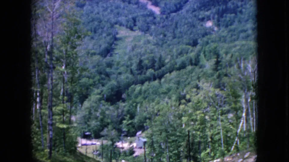 1960s 8mm home-movie view across green wooded hills from a roadside vantage, with the road edge and a small distant structure partly visible.