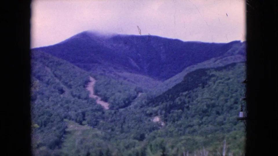 Final 8mm frame of a hazy United States mountain landscape, with forested slopes, a pale winding road or clearing, and dark film-frame edges around the scene.