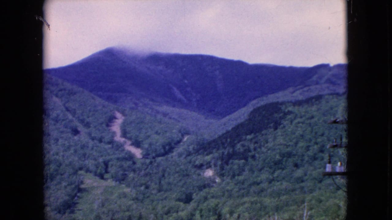 1964 8mm home-movie frame showing a distant forested mountain ridge in the United States, with a pale winding path or cleared run crossing the green hillside and dark structural borders at the sides.