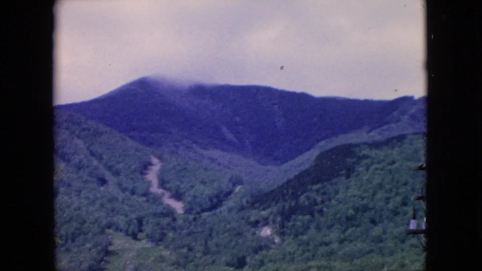 1964 8mm home-movie frame showing a close-framed view of forested hills and a clouded mountain ridge, with a light winding path cutting upward through the valley.
