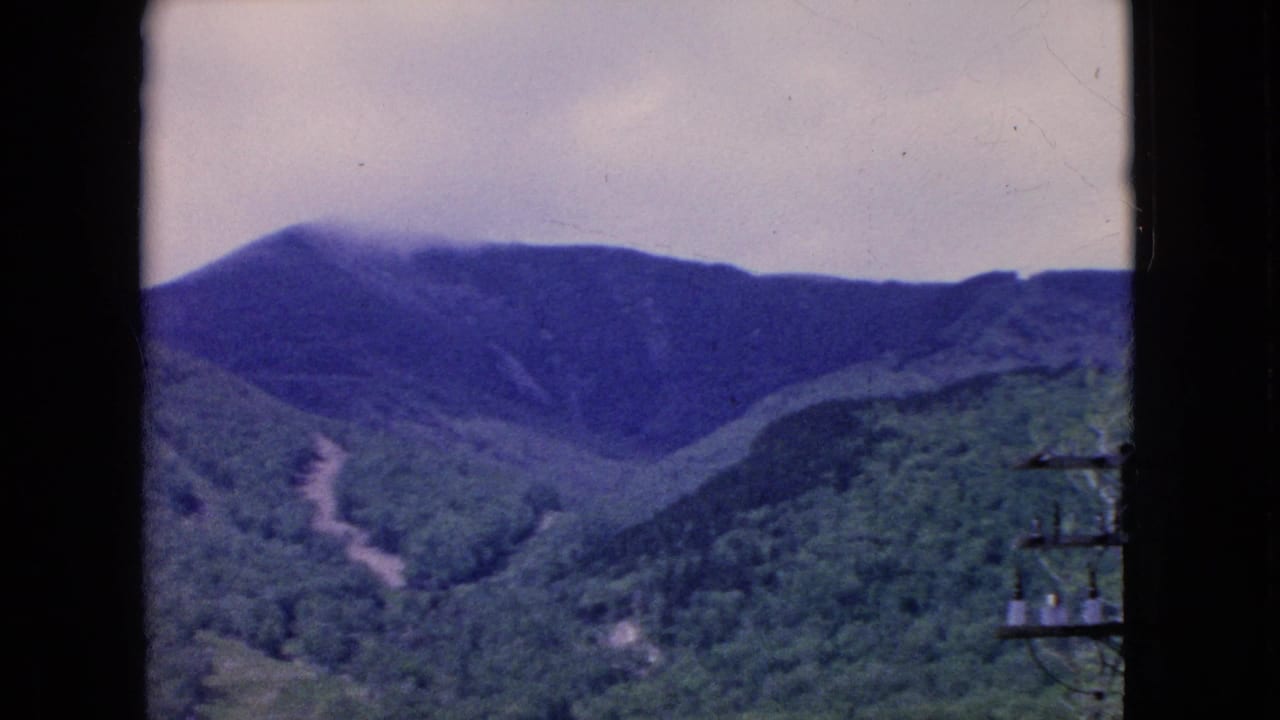 1964 United States 8mm home-movie frame showing a soft, grainy view of a wooded mountain ridge and valley seen through a dark window-like frame, with a utility pole partly visible on the right.