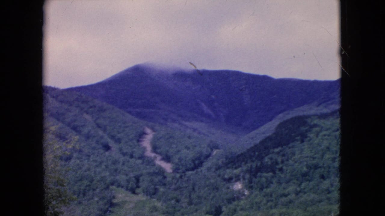 1964 United States 8mm still with a distant mountain ridge softened by mist, green forested slopes below, a pale overcast sky, and dark frame borders from the filmed vantage point.