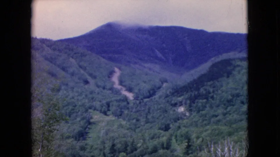1964 United States 8mm still of a green wooded valley beneath a hazy mountain ridge, framed by dark vertical borders with visible grain, scratches, and faded color.
