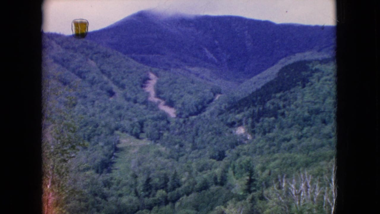 1960s 8mm home-movie frame of a green mountain valley in the United States, viewed through a dark frame with a winding pale road on the hillside.